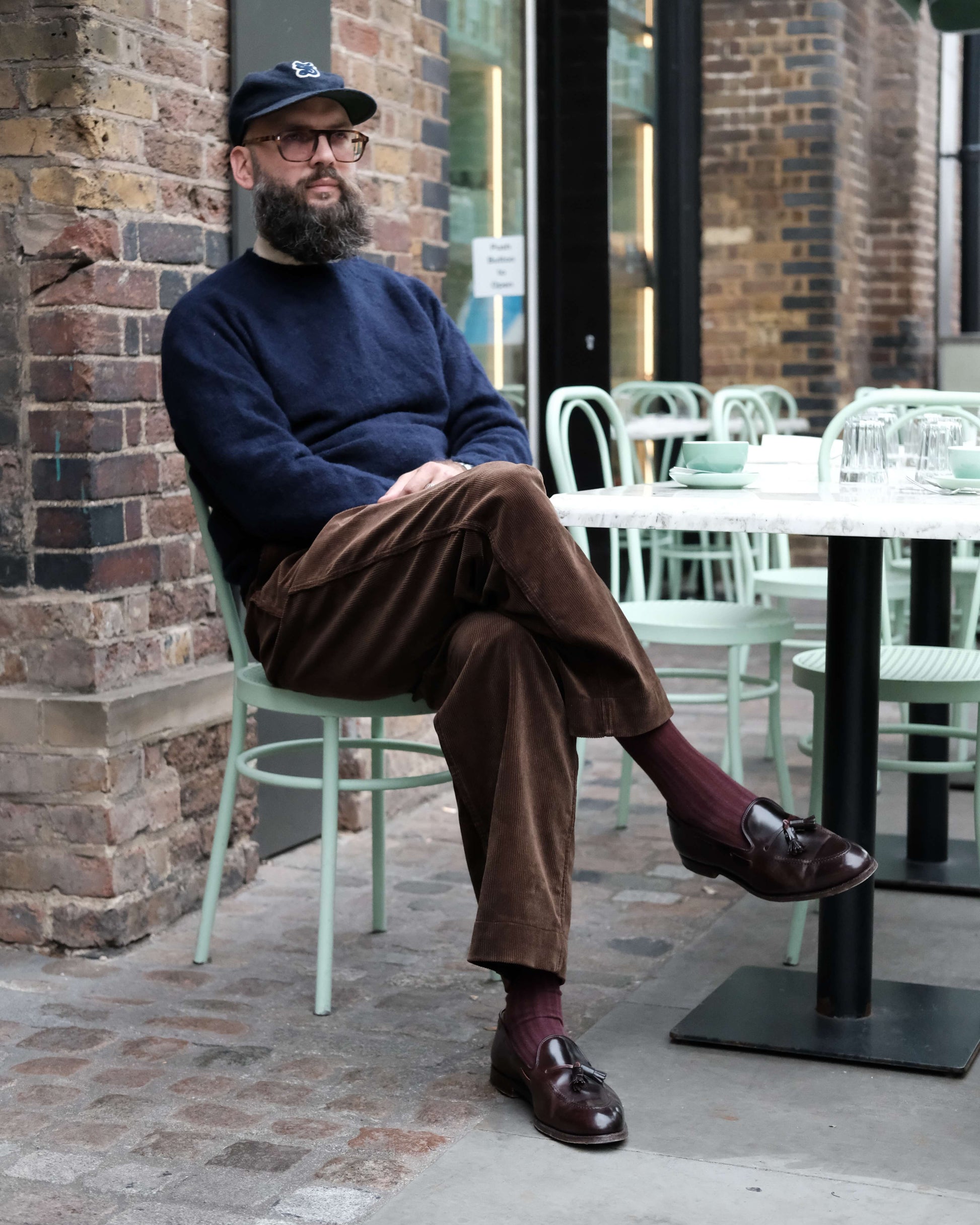 Man sitting on a chair outside a building with a brick wall and tables in the background.